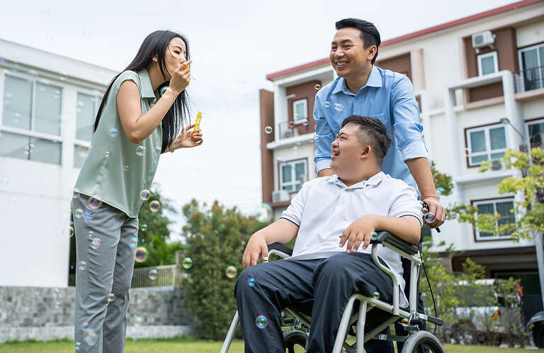 An Asian man and woman enjoy time outside blowing bubbles with and Asian man, who has Down syndrome, watches and smiles from his wheelchair.